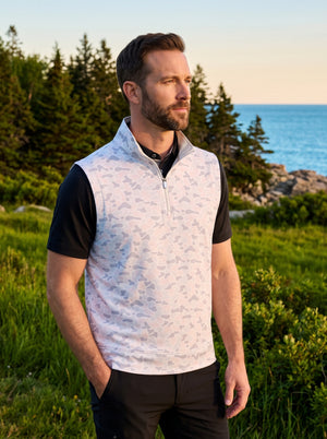 Front upper body angled shot of model wearing brown grey camo full print quarter zip sleeveless vest pullover with one hand in pants pocket and looking into the distance