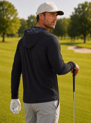Back upper body angled shot of model wearing black soft stretchy hoodie with inner print hood, holding a golf ball in one hand and resting on a golf club with the other hand on the golf course