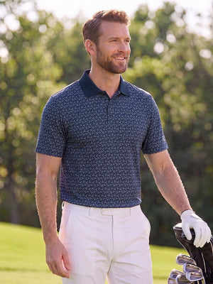 Front upper body angled shot of male model wearing dark blue premium ribbed collar print polo shirt, with one gloved hand resting on golf bag  on the golf course