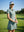 Front shot of model wearing dusty blue women's lightweight golf dress with 2 pockets and hand resting on golf club on the golf course