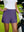 Front mid-length shot of female model wearing dark purple women's gym golf shorts with liner pocket, one hand on waist and one gloved hand resting on golf bag on the golf course